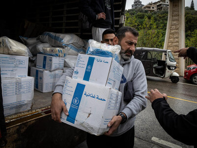 A person carries stacked WFP food‑ration boxes from a truck loaded with supplies, as others assist during a distribution on a roadside.